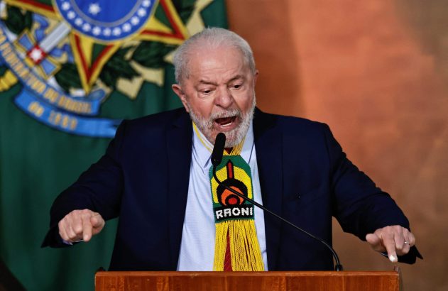 Homem de barba, terno, com colar indígena "RAONI" em púlpito, gesticula e fala. Bandeira do Brasil ao fundo.