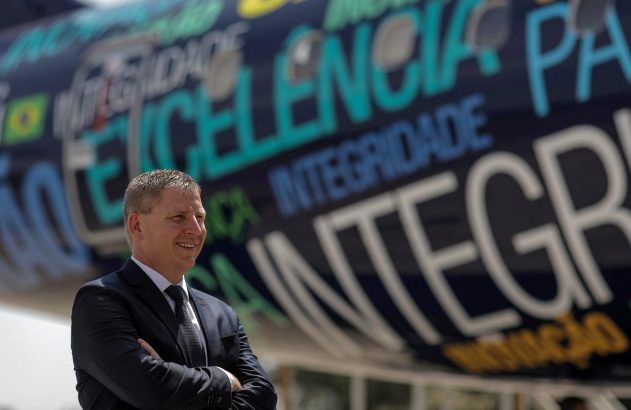 Homem de terno sorrindo, braços cruzados, à frente de avião pintado com palavras como "Excelência" e "Integridade".
