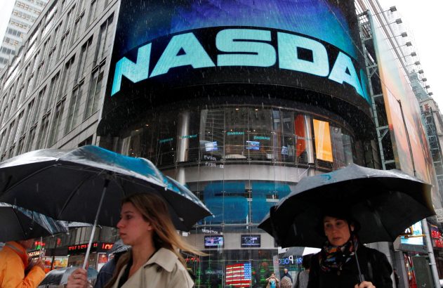 Pessoas com guarda-chuvas caminhando na chuva. Letreiro NASDAQ luminoso no centro urbano.