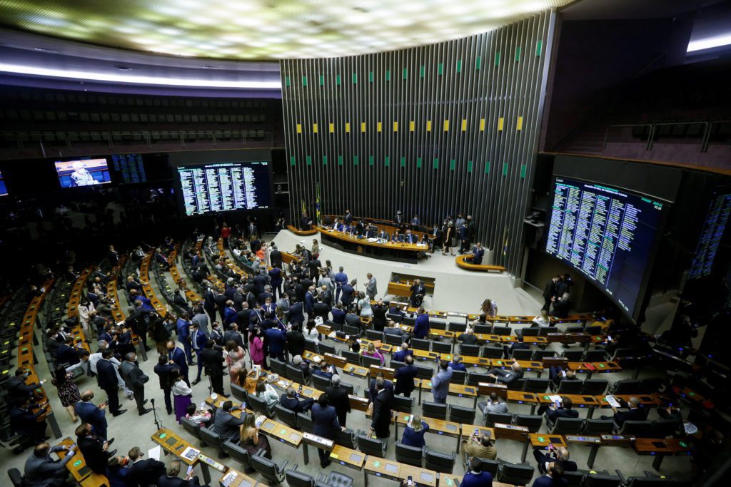 Sessão parlamentar em andamento. Inúmeros políticos em plenário, com telas exibindo informações e dados.