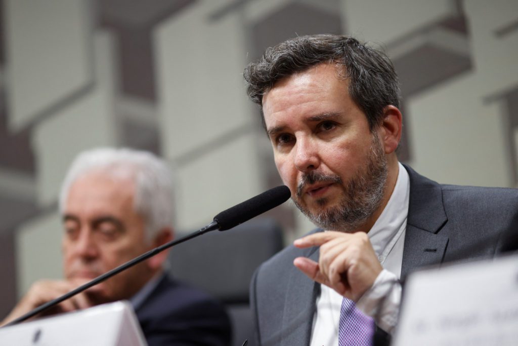 Homem de terno e barba fala ao microfone, gesticulando. Um homem de cabelo branco desfocado está atrás.