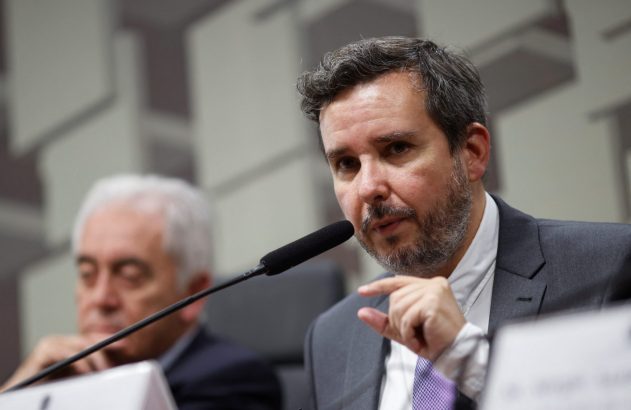 Homem de terno e barba fala ao microfone, gesticulando. Um homem de cabelo branco desfocado está atrás.