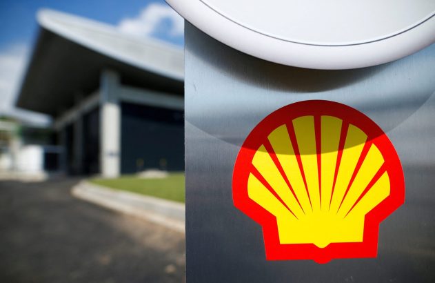 Logo da Shell (concha vermelha e amarela) em superfície metálica. Prédio borrado e céu azul ao fundo.