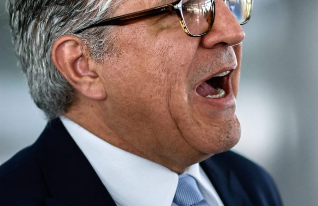 Homem de perfil com cabelo grisalho e óculos, boca aberta como se falasse. Terno escuro, camisa branca e gravata azul.