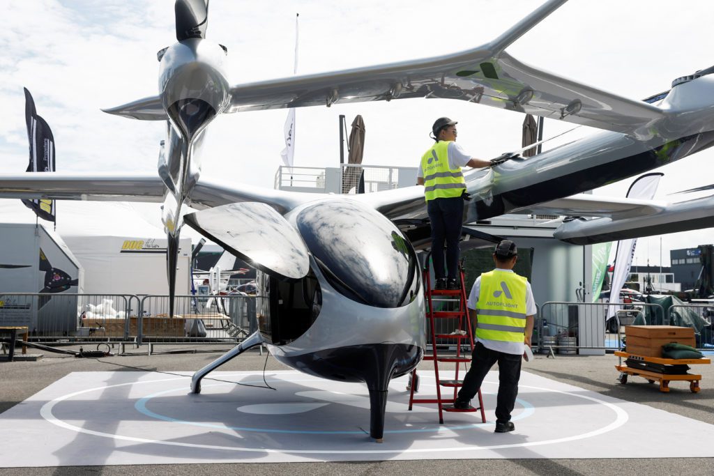 Dois técnicos em coletes Autflight limpam um veículo voador futurista cinza metálico, tipo eVTOL, em uma feira.