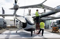 Dois técnicos em coletes Autflight limpam um veículo voador futurista cinza metálico, tipo eVTOL, em uma feira.