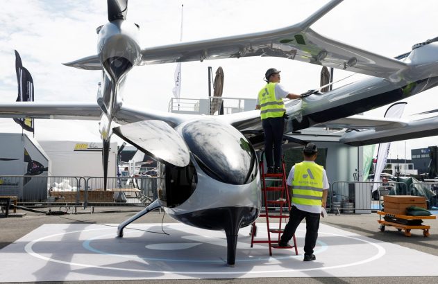 Dois técnicos em coletes Autflight limpam um veículo voador futurista cinza metálico, tipo eVTOL, em uma feira.