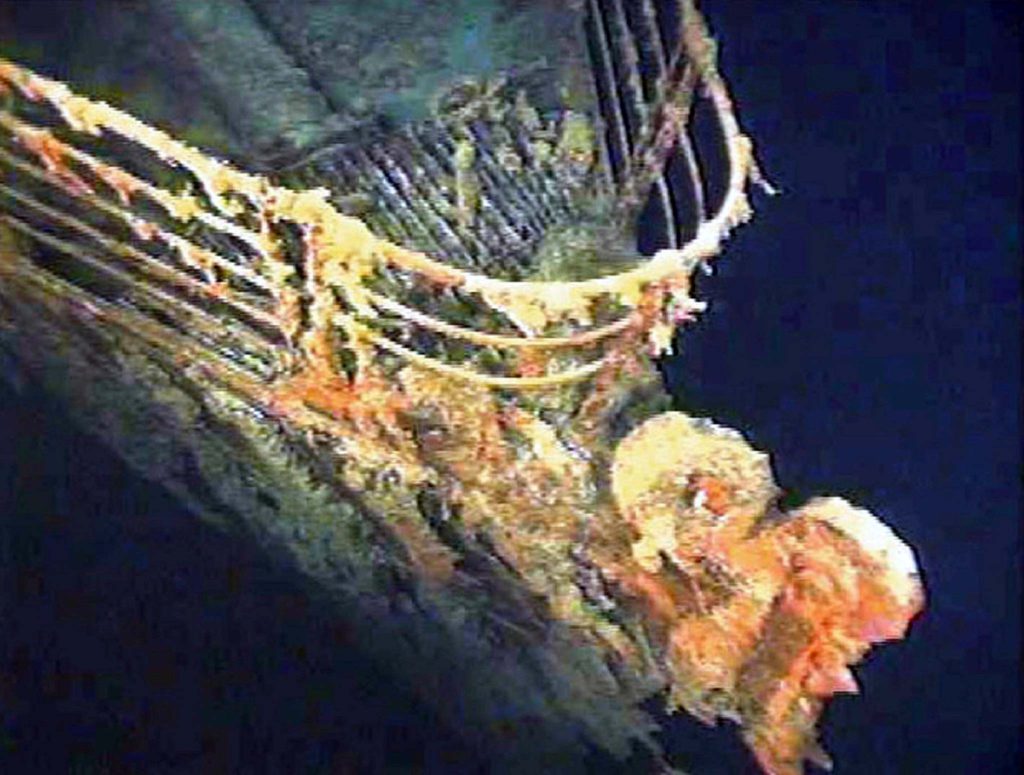 Proa de naufrágio submerso, mostrando corrimões e estrutura corroída por ferrugem. Incrustações alaranjadas e amareladas.