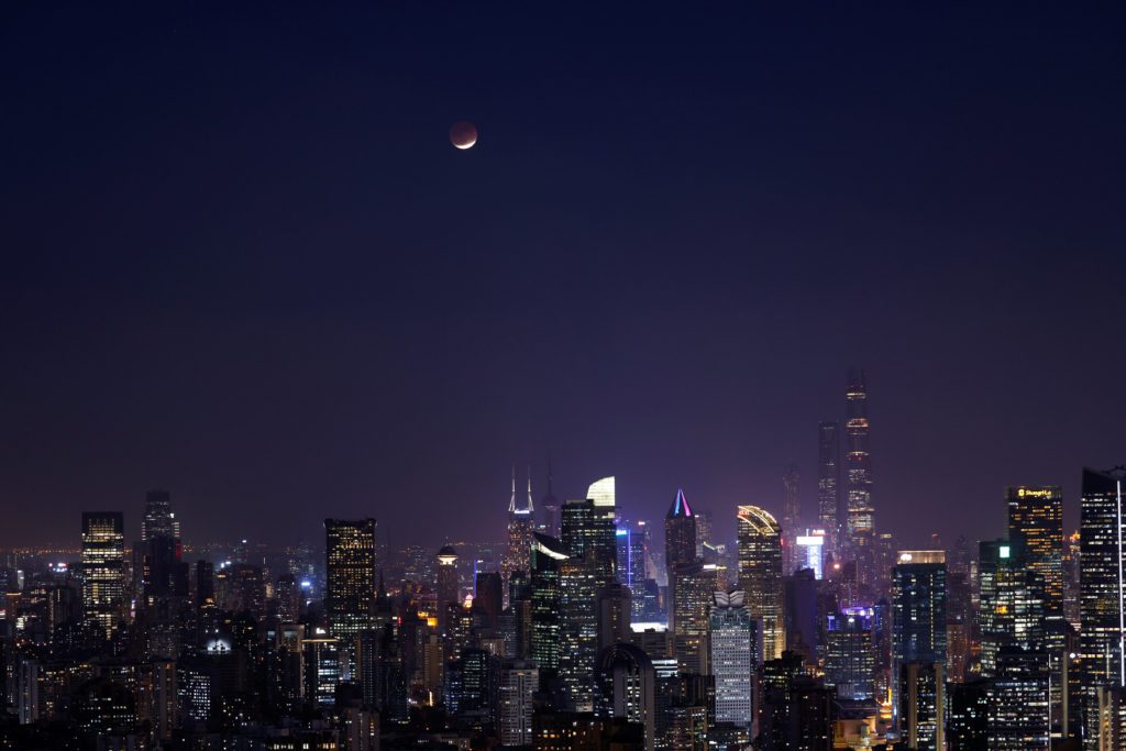 Eclipse lunar avermelhado sobre o skyline noturno de uma cidade iluminada.