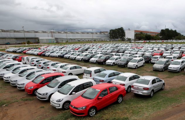 Centenas de carros novos em um amplo estacionamento ao ar livre, com edifícios industriais ao fundo e céu cinzento.