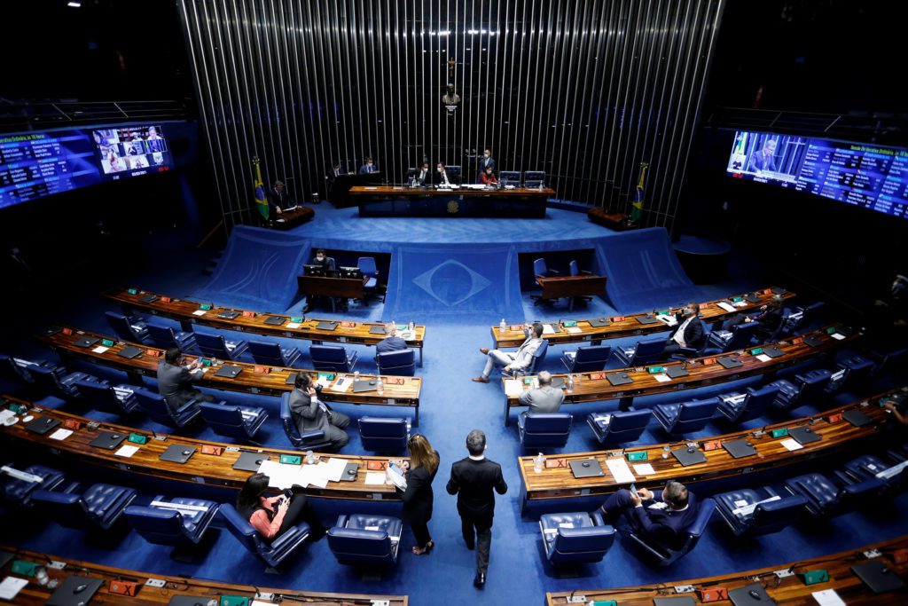 Plenário legislativo com fileiras de assentos azuis. Poucas pessoas caminham no salão, palco e telões ao fundo.