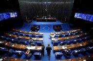 Plenário legislativo com fileiras de assentos azuis. Poucas pessoas caminham no salão, palco e telões ao fundo.