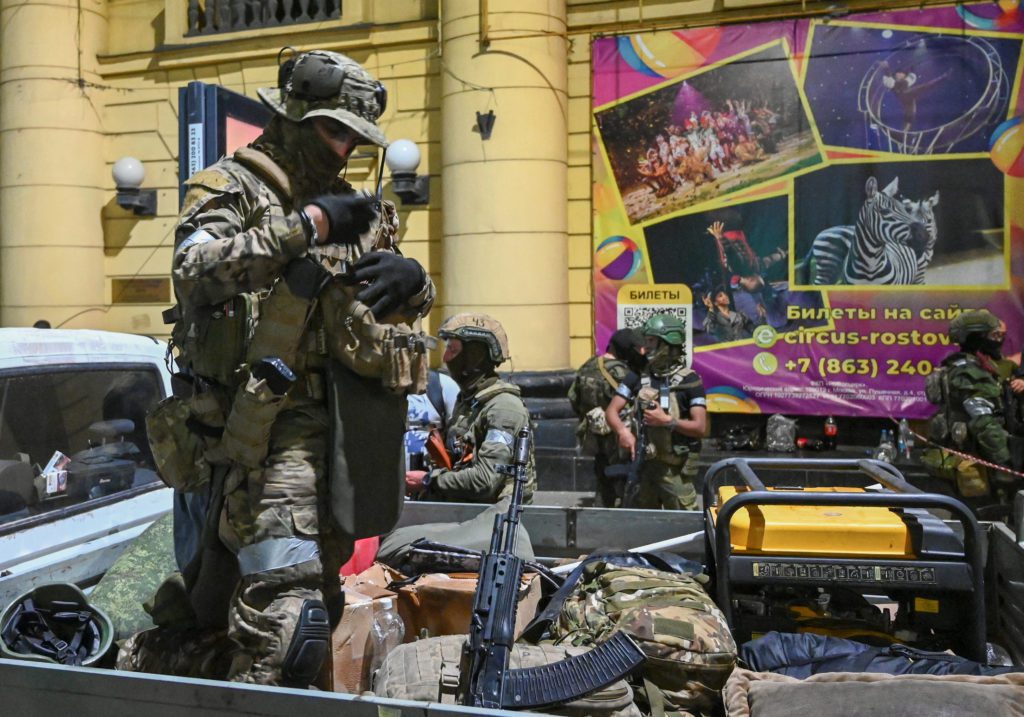 Soldados com equipamento tático e armas ajustam coletes na caçamba de um caminhão, com cartazes de circo ao fundo.