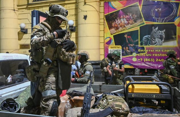 Soldados com equipamento tático e armas ajustam coletes na caçamba de um caminhão, com cartazes de circo ao fundo.