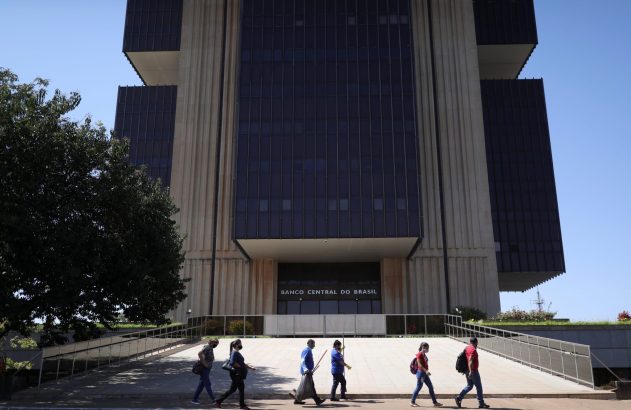 Prédio do Banco Central do Brasil. Pessoas, algumas com máscara, caminham em frente sob céu azul.