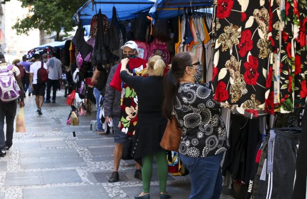 Pessoas em feira de rua movimentada olhando roupas em barracas, algumas com máscara.