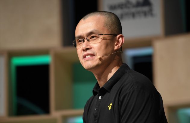 Changpeng Zhao (CZ) com óculos e microfone, em camisa polo preta com logo Binance, falando em evento.