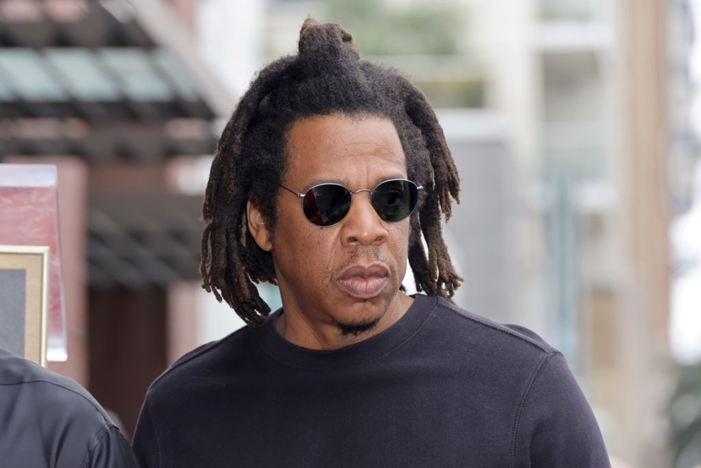 Jay-Z, com dreadlocks e óculos de sol redondos, sério, olhando para o lado. Fundo urbano.