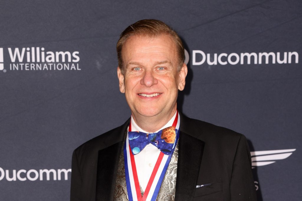 Retrato de homem sorridente em smoking com gravata borboleta de planetas e medalha. Fundos: Williams e Ducommun.
