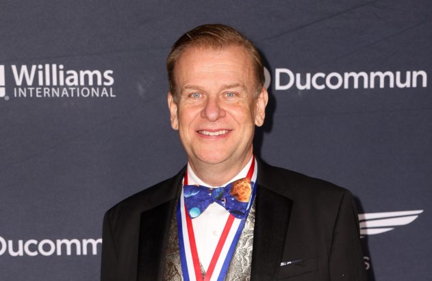 Retrato de homem sorridente em smoking com gravata borboleta de planetas e medalha. Fundos: Williams e Ducommun.