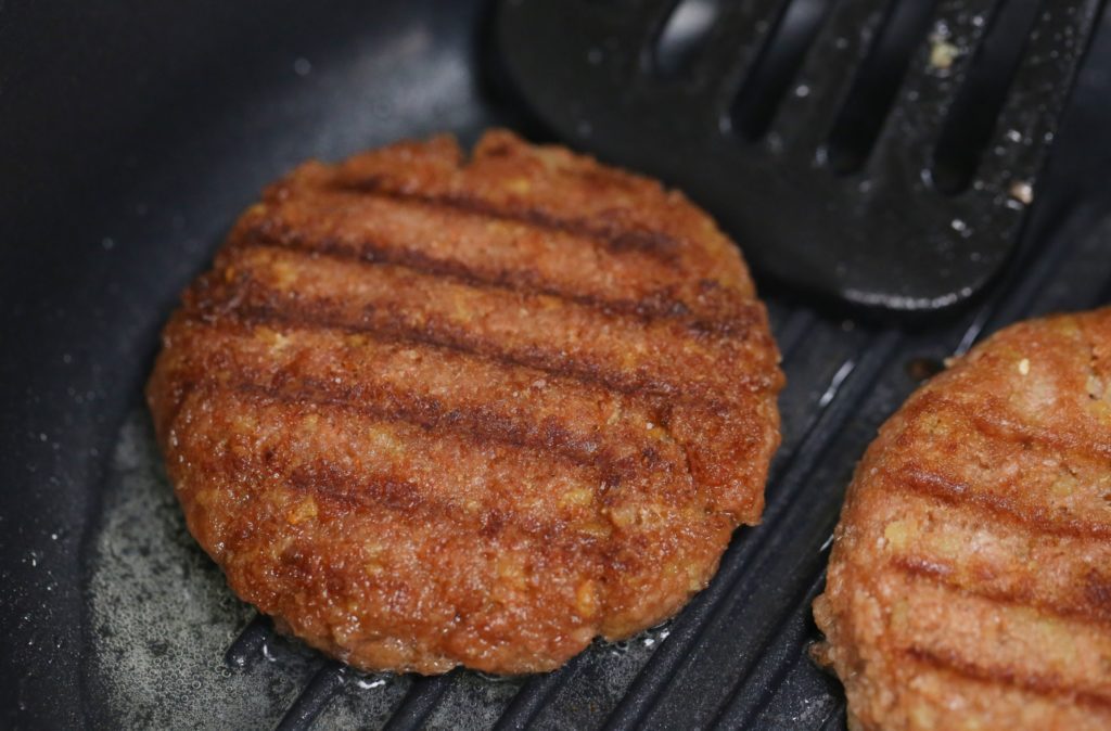 Hambúrgueres com marcas de grelha cozinhando em chapa preta. Óleo borbulhando; espátula visível.