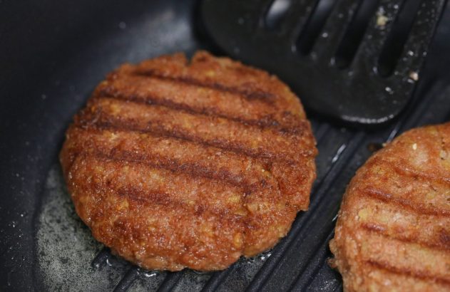 Hambúrgueres com marcas de grelha cozinhando em chapa preta. Óleo borbulhando; espátula visível.