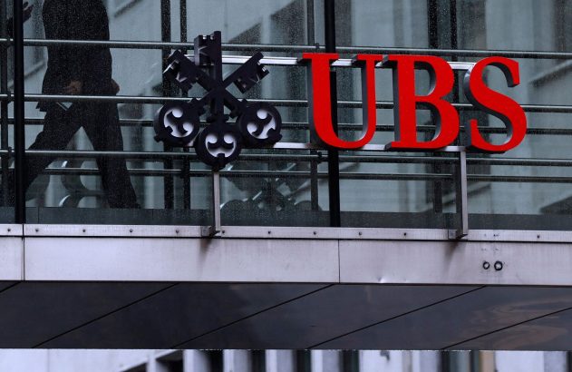 Logo vermelho da UBS e símbolo das chaves em prédio. Silhuetas de pessoas em passarela superior.