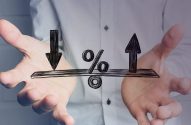 Mãos seguram balança com seta para baixo, porcentagem e seta para cima. Simboliza equilíbrio ou variação de taxas.