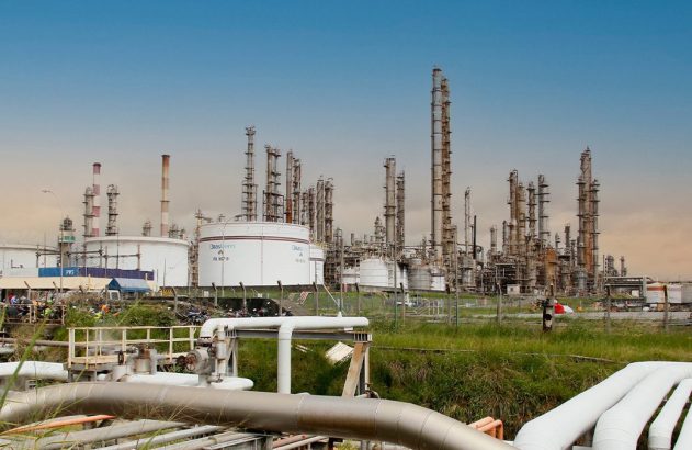 Refinaria de petróleo com torres, tanques e tubulações em paisagem industrial sob céu azul.