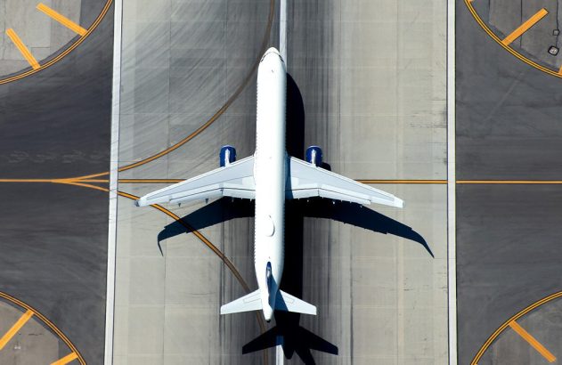 Vista aérea de avião branco com motores azuis em pátio de aeroporto. Há marcações amarelas no asfalto.