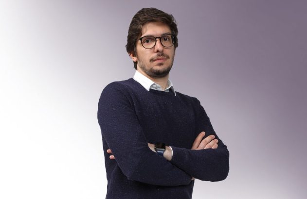 Homem de óculos, barba e suéter azul, com braços cruzados.