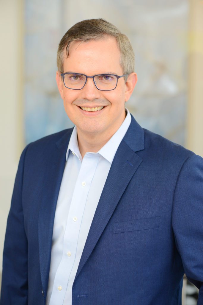 Retrato de um homem sorridente de óculos, vestindo blazer azul e camisa clara.