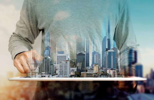 Mãos seguram modelo de cidade em tablet. Ao fundo, paisagem urbana com céu azul.