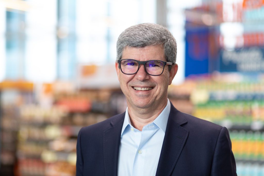 Homem sorrindo, usando óculos e paletó, em supermercado.