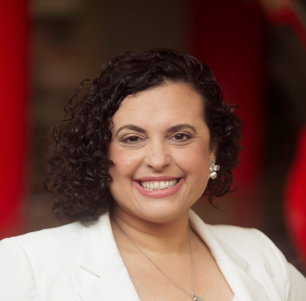 Mulher sorrindo, cabelo escuro cacheado, blazer branco, colar, contra fundo vermelho desfocado.