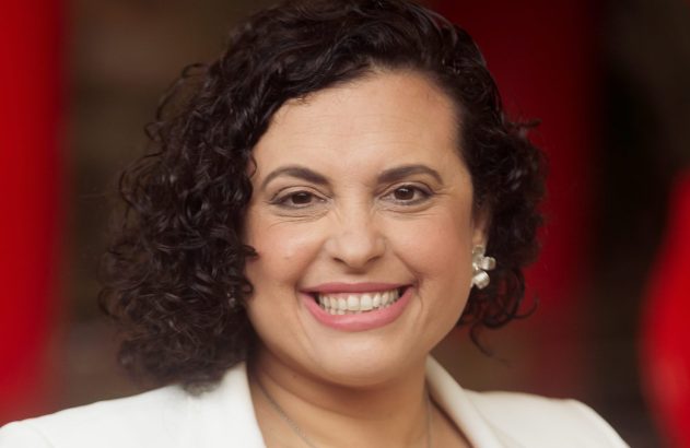 Mulher sorrindo, cabelo escuro cacheado, blazer branco, colar, contra fundo vermelho desfocado.