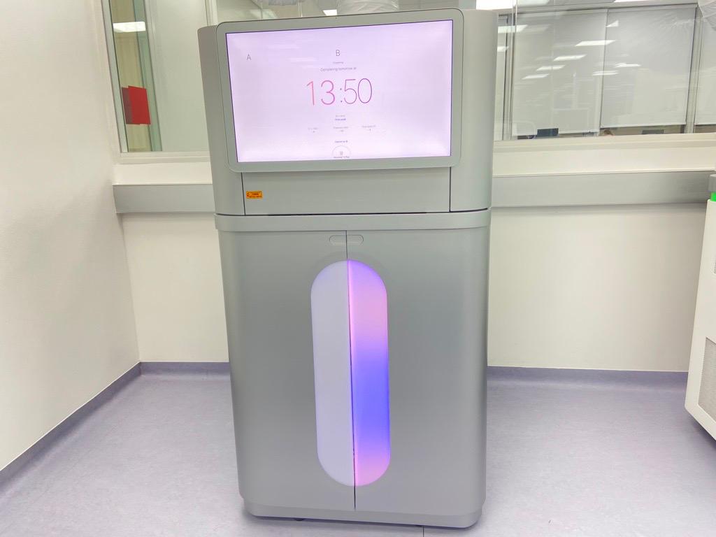 Máquina cinza em laboratório, com tela mostrando "13:00" e painel vertical iluminado em roxo.