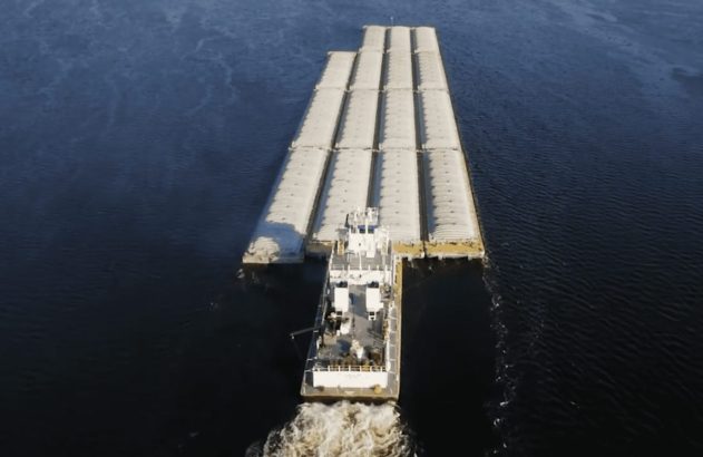 Vista aérea de um empurrador empurrando um comboio de grandes barcaças carregadas em um rio.