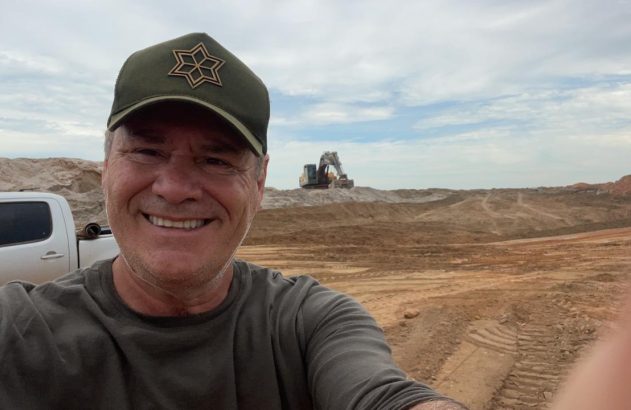 Homem sorrindo de boné verde com estrela tira selfie em local de terraplanagem com caminhonete e escavadeira.