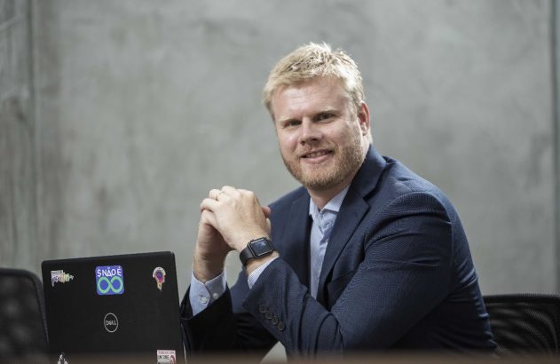 Homem sorrindo com barba e blazer azul, sentado em mesa com as mãos unidas e laptop à frente.