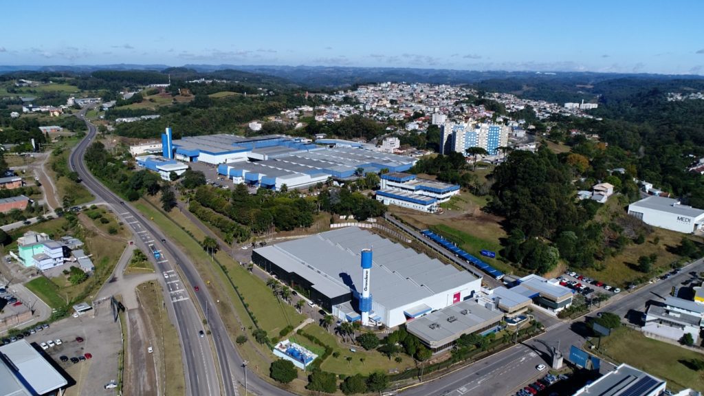 Vista aérea: parque industrial com galpões azuis, rodovia e cidade em dia ensolarado.