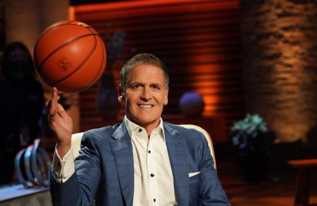 Mark Cuban sorrindo, girando uma bola de basquete no dedo. Ele usa um terno e está sentado em um cenário de TV.