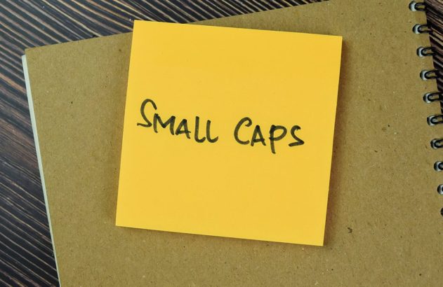 Post-it amarelo "Small Caps" em caderno espiral marrom sobre mesa de madeira.