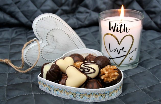 Caixa de chocolates em formato de coração, coração decorativo e vela acesa com 'With Love' sobre tecido cinza acolchoado.