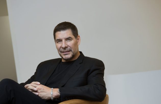 Retrato de homem de terno preto, sentado com mãos cruzadas e pulseira, olhando para a câmera.