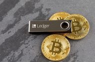 Uma carteira de hardware Ledger, em forma de pen drive, e três moedas douradas de Bitcoin.