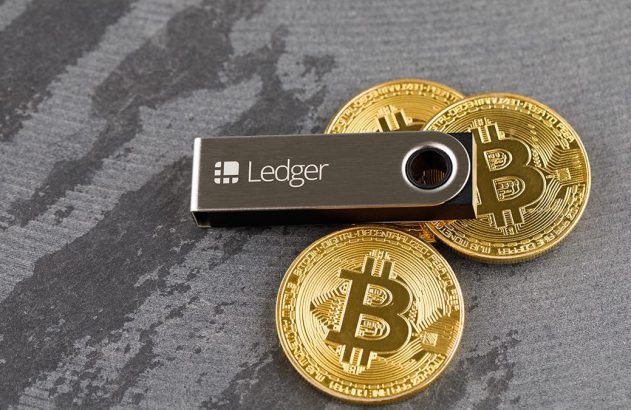 Uma carteira de hardware Ledger, em forma de pen drive, e três moedas douradas de Bitcoin.