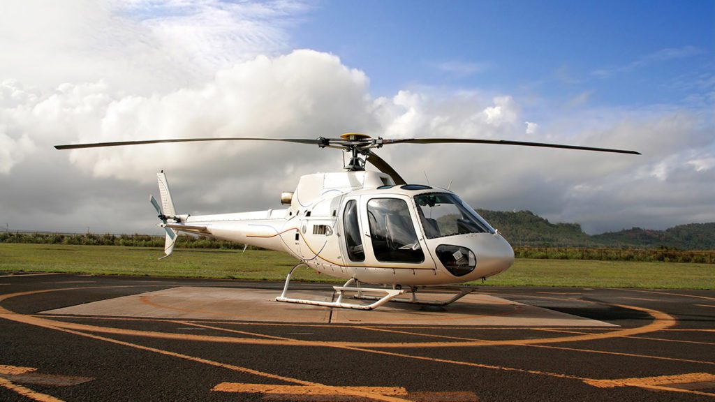 Helicóptero branco no heliponto, gramado e montanhas distantes sob céu parcialmente nublado.