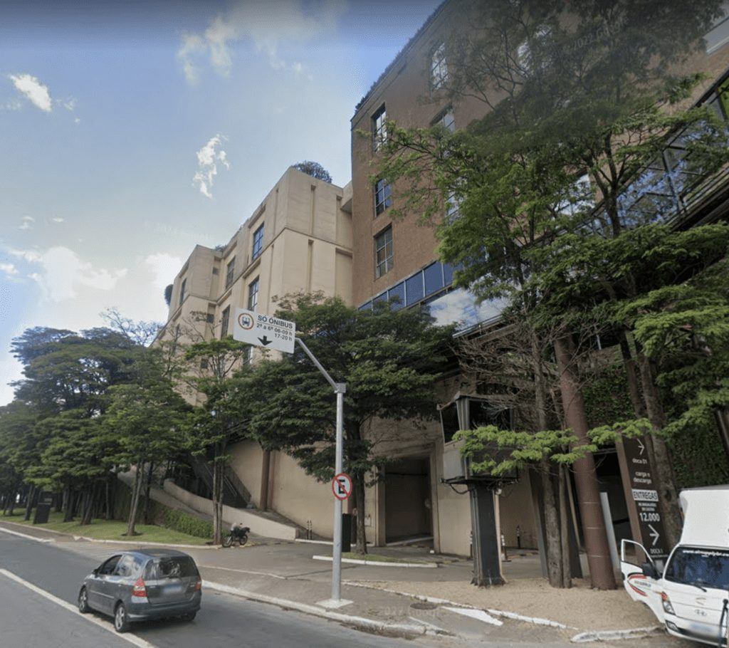 Cena urbana: Carro na rua de dia, com edifício em declive coberto por árvores ao fundo. Céu azul claro.