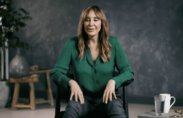 Mulher de cabelos castanhos, blusa verde e calça escura, sentada numa cadeira preta, com as mãos nas pernas e sorriso discreto. Fundo neutro.